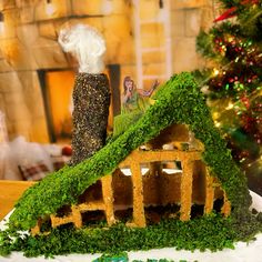 a house made out of moss with a doll in the back ground and a christmas tree behind it