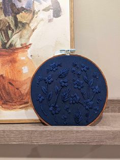 an embroidered blue piece of art next to a vase with flowers on it and a painting in the background