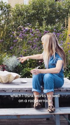 a woman is sitting on a bench in front of some flowers and holding her hand out