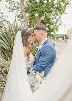 a bride and groom standing together in front of some flowers at their wedding day, looking into each other's eyes