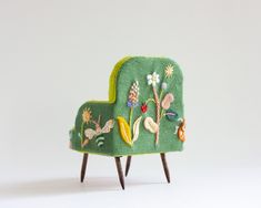 a small green chair with flowers and plants on it's back, sitting in front of a white wall