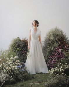 a woman in a white dress standing on top of a lush green field next to flowers