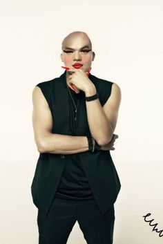 a man in a black vest and red lipstick holding his hand to his mouth while standing against a white wall