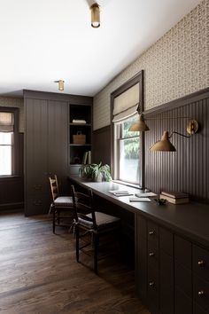 a desk and chair in a room with dark wood paneling on the walls, along with two windows