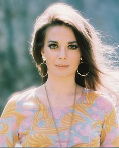 a woman with long hair and large hoop earrings standing in front of a wall wearing a colorful top