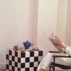 a woman sitting in a chair reading a book while holding a cup and saucer