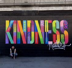 a man sitting on the ground in front of a wall that has been painted with colorful letters