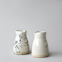 two white vases sitting side by side on a gray surface, one with black speckles