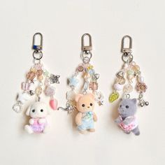 three key chains with small stuffed animals hanging from them on a white surface, one is holding a teddy bear and the other has charms attached to it's ear