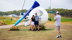 three people are standing in front of a golf ball and tee on the grass, with one person taking a photo