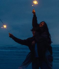 two people holding sparklers in their hands on the beach at night with dark sky and ocean behind them