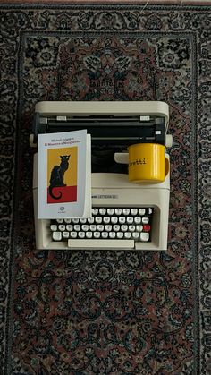 an old fashioned typewriter sitting on top of a rug next to a yellow cup