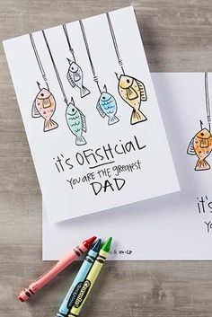 two greeting cards with colored pencils next to them on a wooden surface, one has an image of fish and the other says it's official you are the first dad