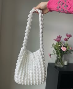 a hand holding a white crocheted purse next to a vase with pink flowers