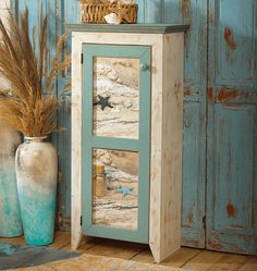 a blue cabinet with two doors and starfishs on the glass door is next to a vase with dried grass in it