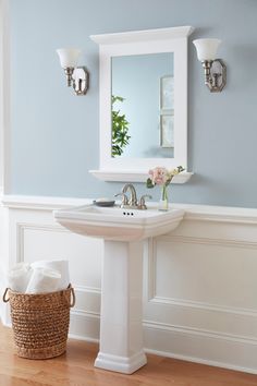 a white pedestal sink sitting under a mirror next to a basket with flowers on it