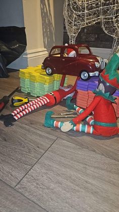 the elf is sitting on the floor with his toy car and stack of gifts in front of him