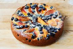 a cake with blueberries, peaches and powdered sugar sits on a wooden table