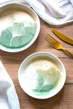 two white bowls filled with green liquid next to gold utensils on top of a wooden table