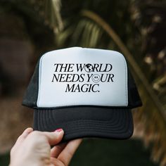 a person holding up a black and white trucker hat that says the world needs your magic