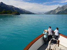 three people standing on the bow of a boat looking out at mountains and blue water