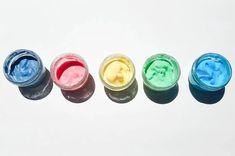 four jars filled with different colored powders on top of a white surface and one is empty