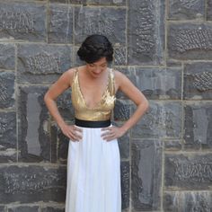 a woman standing in front of a stone wall wearing a white and gold dress with her hands on her hips