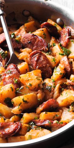 a pan filled with sausage and potatoes on top of a wooden table next to a fork