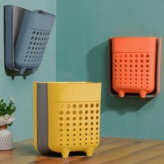 two orange and one blue baskets are on the wall next to each other in front of a potted plant