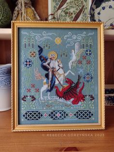 a cross stitch picture in a gold frame on a wooden table next to some vases