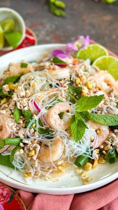 a white plate topped with noodles and shrimp next to lime wedges on a pink cloth