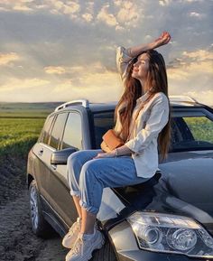 a woman sitting on the hood of a car in front of a field and sky