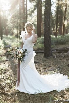 a woman in a white wedding dress standing in the woods with her bouquet on her shoulder