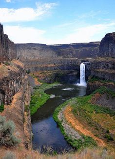 a large waterfall in the middle of a canyon with green grass and bushes around it