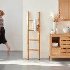 a woman walking past a bathroom sink next to a wooden cabinet and ladder with towels on it