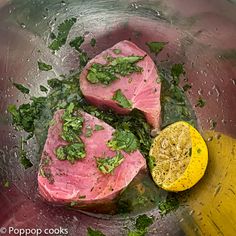 two pieces of raw meat in a bowl with herbs on top and lemon wedges