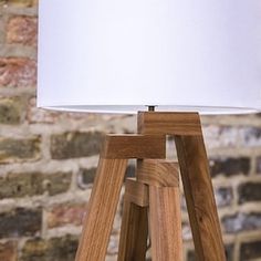 a wooden tripod table lamp with a white shade on it in front of a brick wall