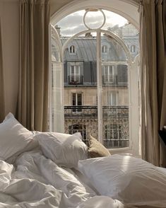 an unmade bed with white sheets and pillows in front of a large arched window