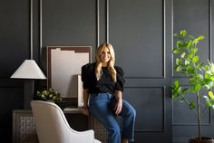 a woman sitting on a white chair in front of a gray wall and potted plant