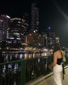 a woman is standing on a bridge looking at the city lights in the night time