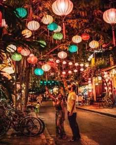 two people standing in the middle of a street with many lanterns hanging from the ceiling