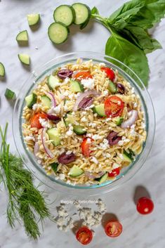 a glass bowl filled with pasta salad next to cucumbers, tomatoes and parsley