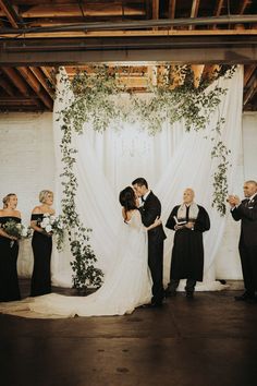 a bride and groom kissing in front of their wedding ceremony arch decorated with greenery