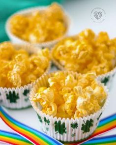 three small cups filled with yellow popcorn on top of a rainbow colored table cloth next to other cupcakes