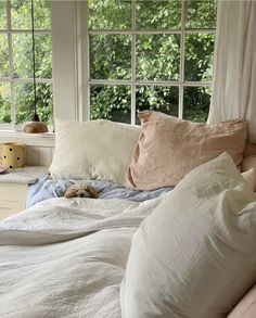 an unmade bed in front of two windows with white curtains and pillows on it