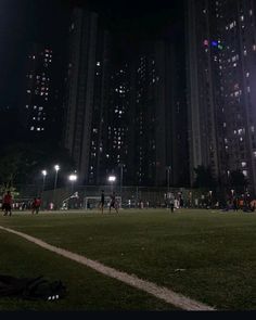 people are playing soccer at night in the city with tall buildings behind them and lights shining on the grass
