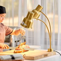 a woman cutting pizza on top of a wooden cutting board next to a metal lamp