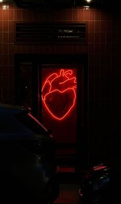 a red neon heart sign on the side of a building next to two parked cars