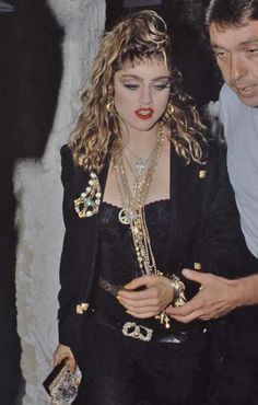 a man standing next to a woman in a black dress and gold chains on her neck