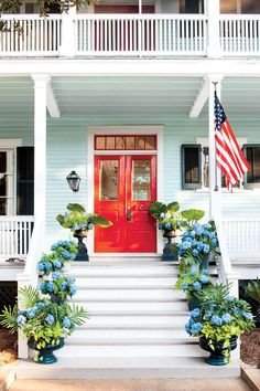 a red door sits on the front steps of a white house with blue hydrangeas and potted plants